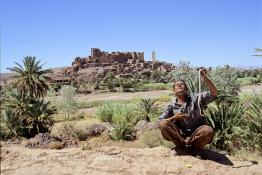 Image du Maroc Professionnelle de  Le paysage, formé par la kasbah Tifoultoute qui se dresse majestueusement sur un éperon rocheux qui domine la vallée de l'oued Ouarzazate, attire les touristes qui prennent le temps de s'arrêter pour prendre une photo. Précisément à cet endroit un charmeur de serpent tenant une vipère avec sa main gauche tout en surveillant le mouvement du scorpion noir qu’il avait accroché à son cou avec sa main droite pose aux touristes contre un billet avec derrière lui comme décor celui qui avait servit pour de grand films comme Lawrence d’Arabie et Jésus de Nazareth à quelque kilomètres de la ville de Ouarzazate au sud du Maroc le Samedi 23 Août 1997. (Photo / Abdeljalil Bounhar) 